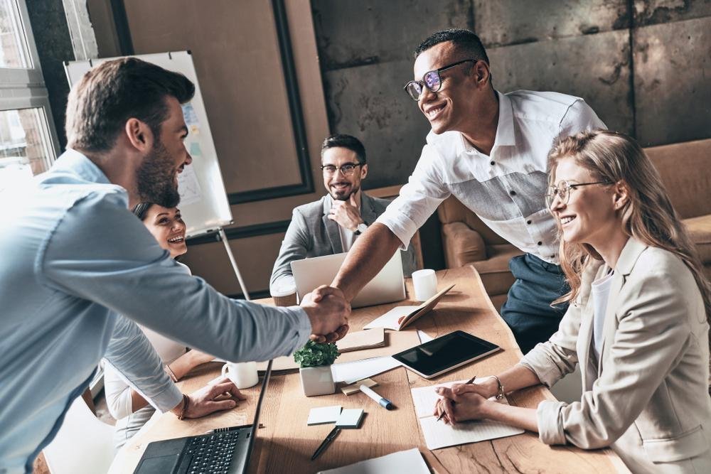 A round table meeting. Two people are shaking hands. All are dressed casually.